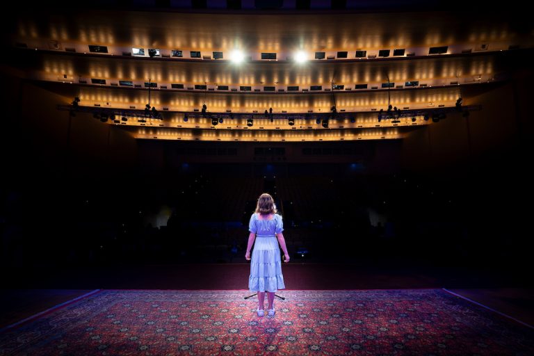 A student stands alone on a stage looking out into an auditorium.